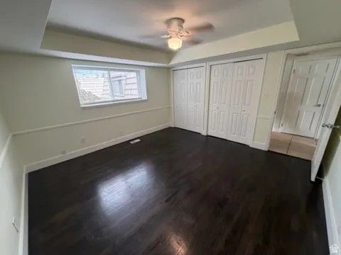 Dark wood-finish flooring, tray ceiling, ceiling fan with integrated lighting, window with blinds, and white bi-fold closet doors