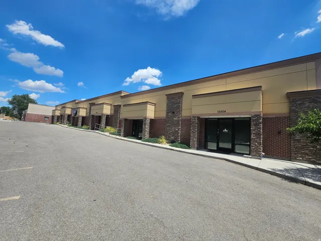 Commercial building exterior featuring extensive asphalt paving, multiple storefronts with glass entry doors, brick lower facade, stone column accents, and a light-colored upper facade
