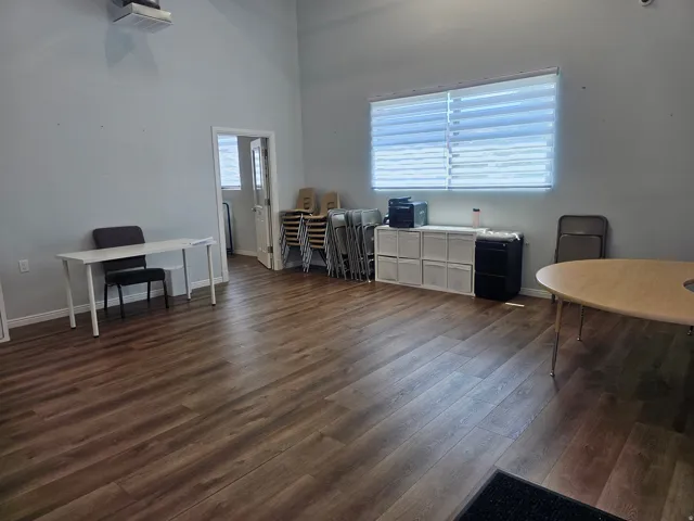 Wood-finish flooring, high vaulted ceiling, large windows with blinds, light-toned wall paint, and a white interior door frame