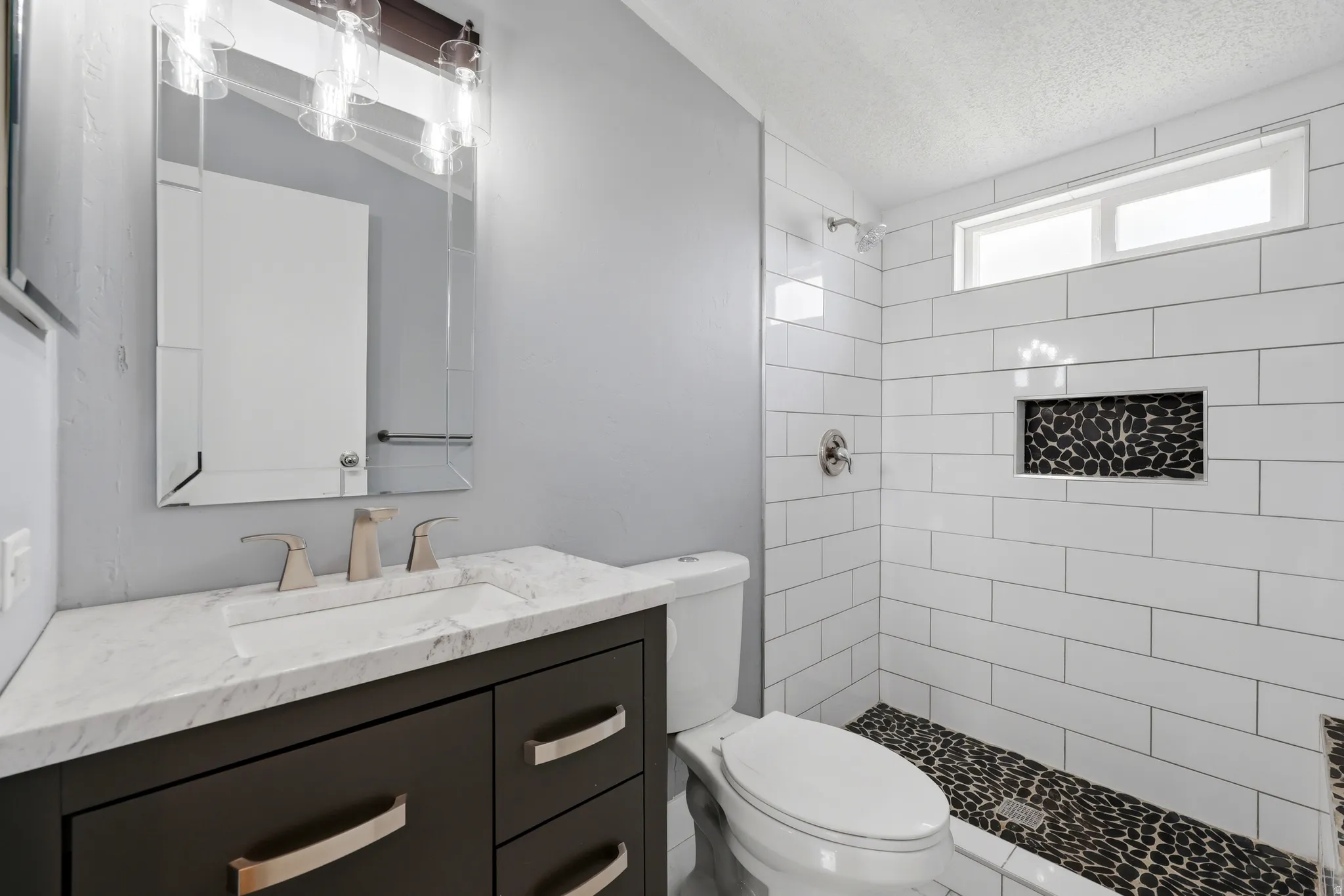 Bathroom featuring a dark wood-finish vanity with a light-toned countertop, a frameless vanity mirror, a tiled shower with a black pebble-style floor, and a horizontal window