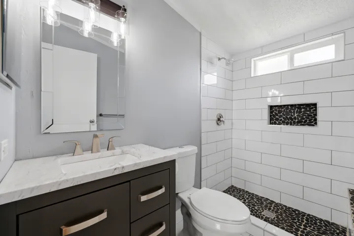 Bathroom featuring a dark wood-finish vanity with a light-toned countertop, a frameless vanity mirror, a tiled shower with a black pebble-style floor, and a horizontal window