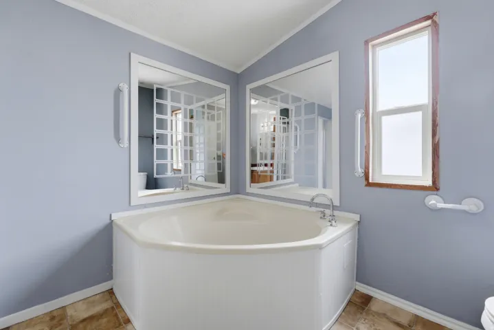 Corner soaking tub with chrome fixtures, two wall-mounted mirrors, wood-frame window, and a tile-finish floor
