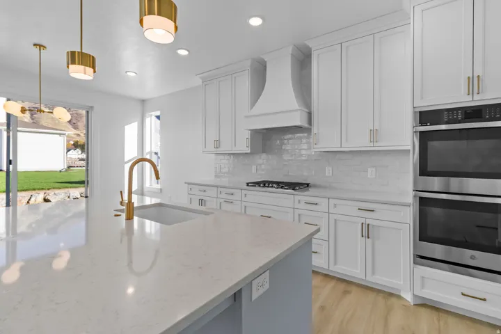 White shaker cabinetry, light-toned countertops, a gold-finish gooseneck faucet, a built-in double oven, and wood-finish flooring