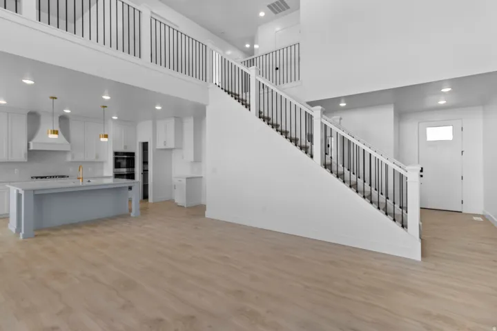 Expansive open-concept living space with wood-finish flooring, a contemporary kitchen featuring a light gray island, white cabinetry, and stainless steel appliances, and a grand staircase with dark metal balusters
