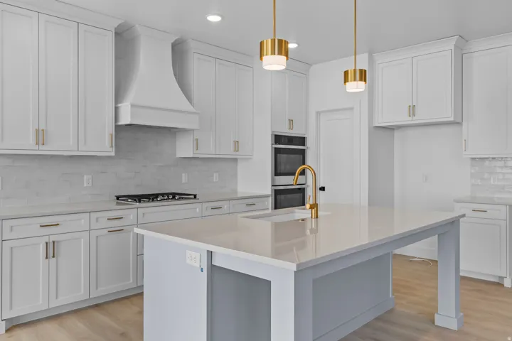 Spacious kitchen featuring white shaker cabinetry, light-toned countertops, a central island with an integrated sink, built-in double ovens, and gold-tone fixtures