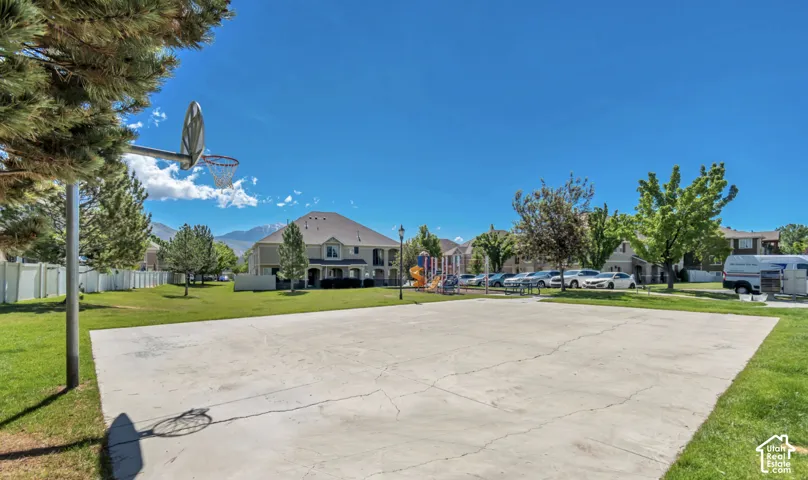 Paved court with an attached basketball hoop, surrounded by green space, a white privacy fence, and distant mountains
