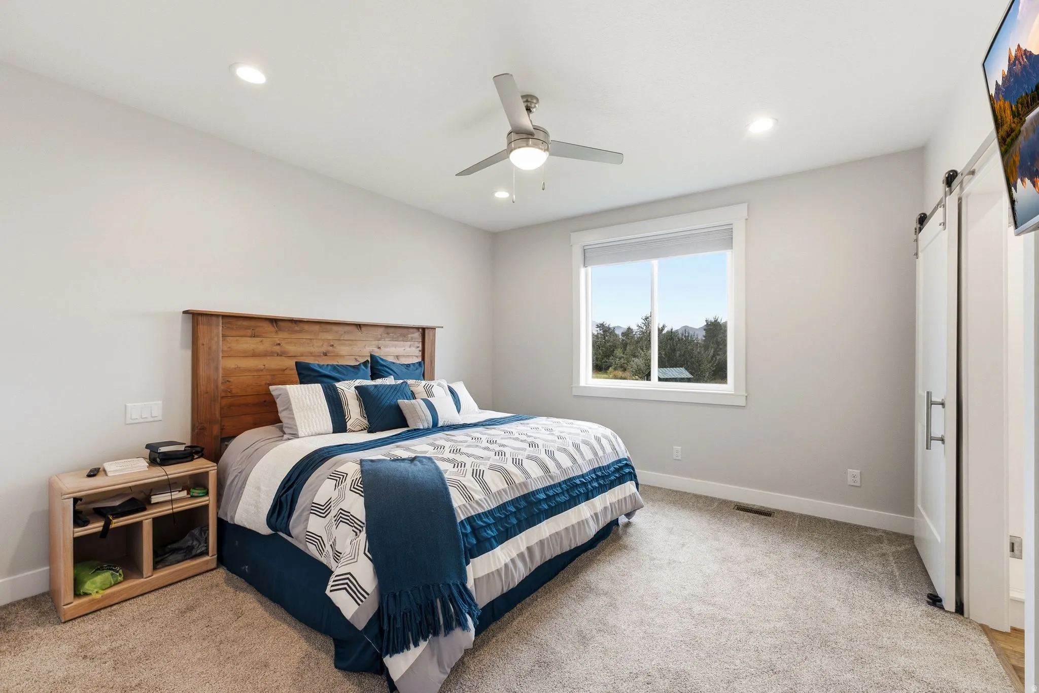 Recessed lighting, ceiling fan, wood-finish headboard, window with outdoor scenery, and barn door
