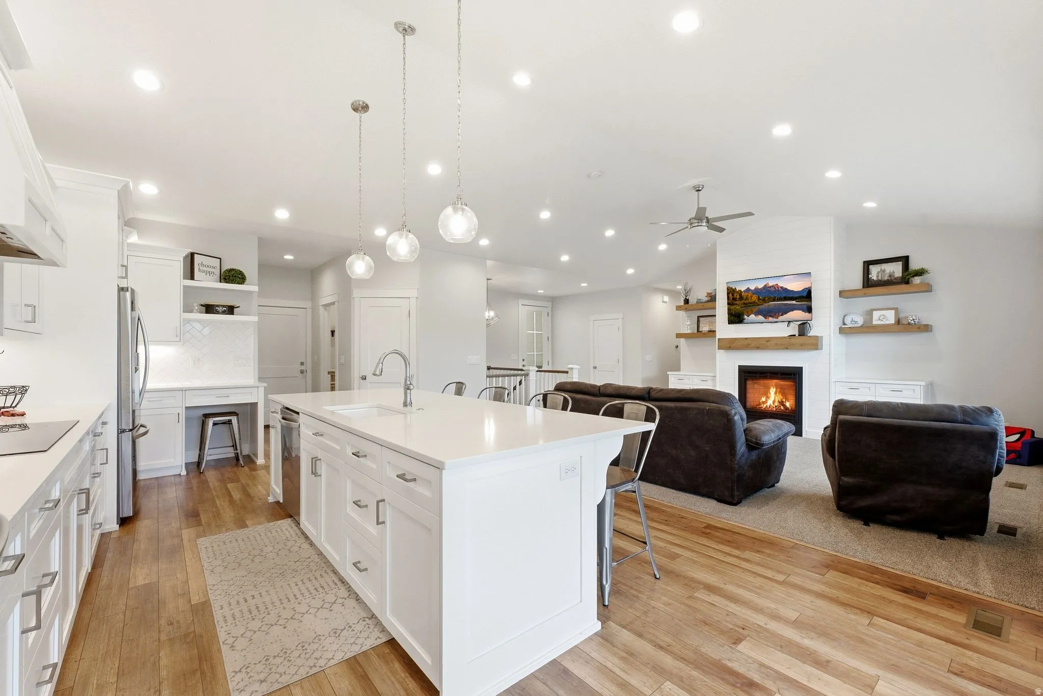 Open-concept kitchen and living area featuring wood-finish flooring, white cabinetry, a kitchen island with seating, a built-in desk, and a fireplace with a wood mantle