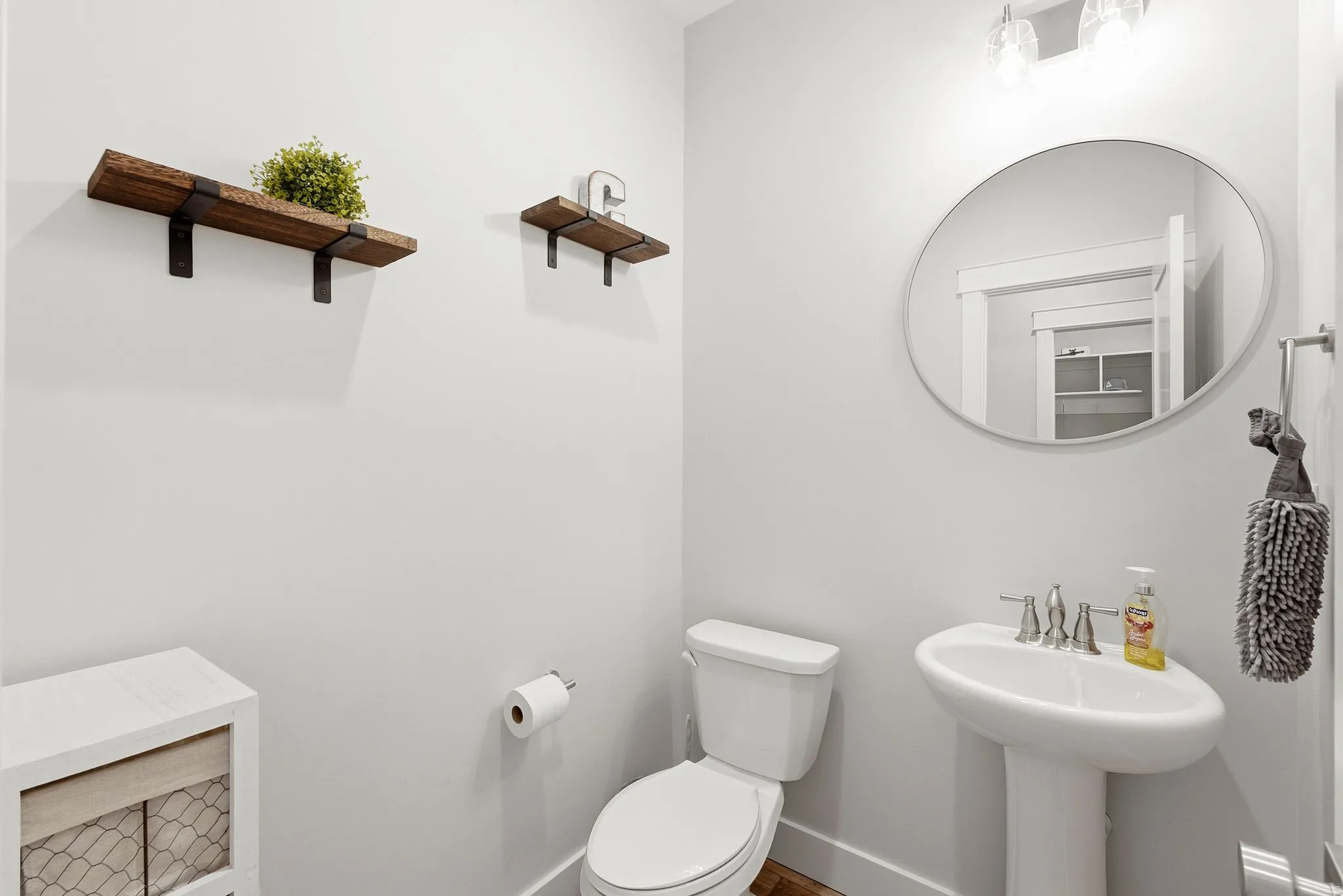 Pedestal sink with a brushed nickel faucet, round mirror, overhead vanity lighting, and wall-mounted shelving