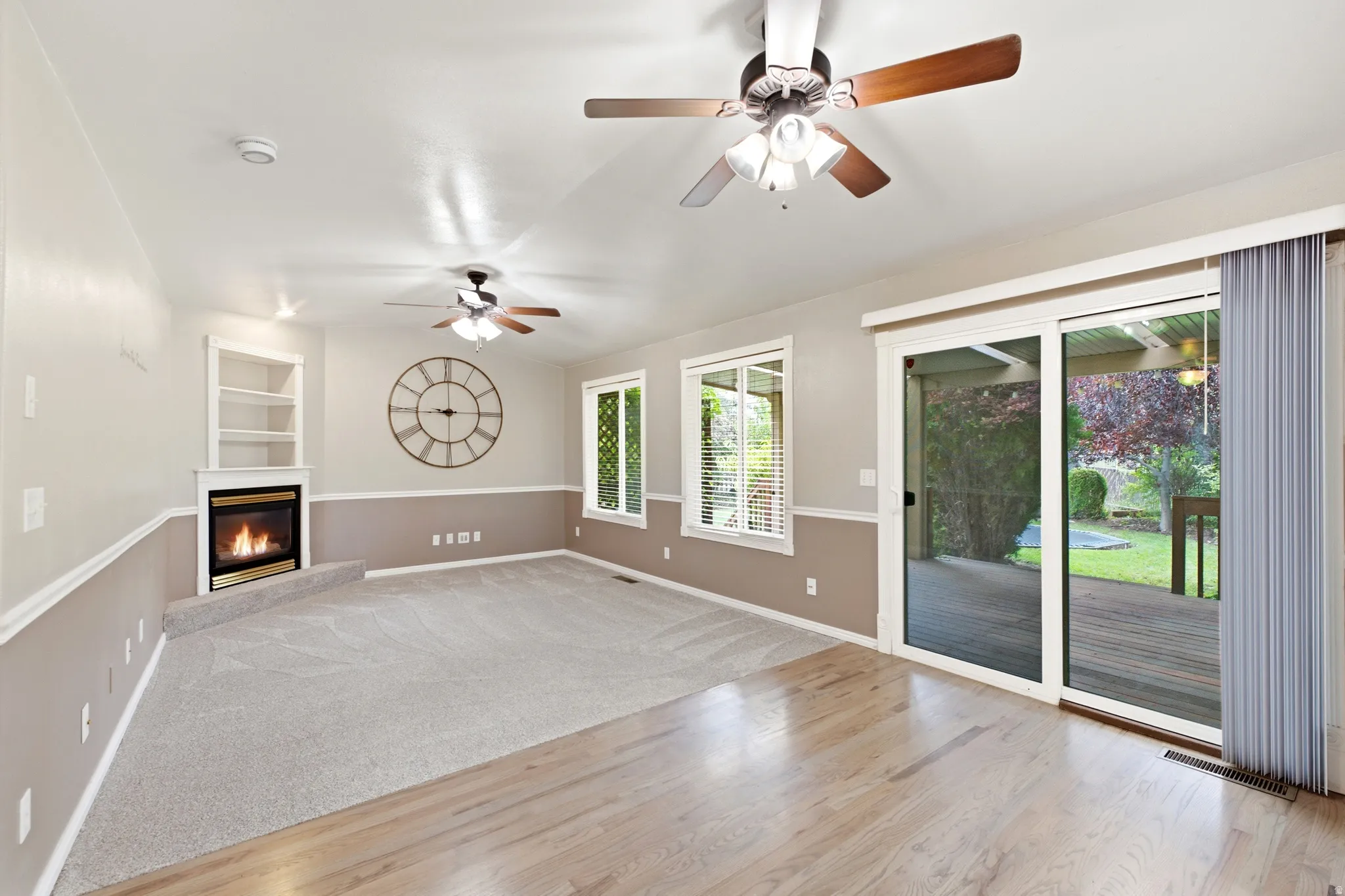 Fireplace with built-in shelving, two ceiling fans, wood-finish flooring, sliding glass door, and backyard decking