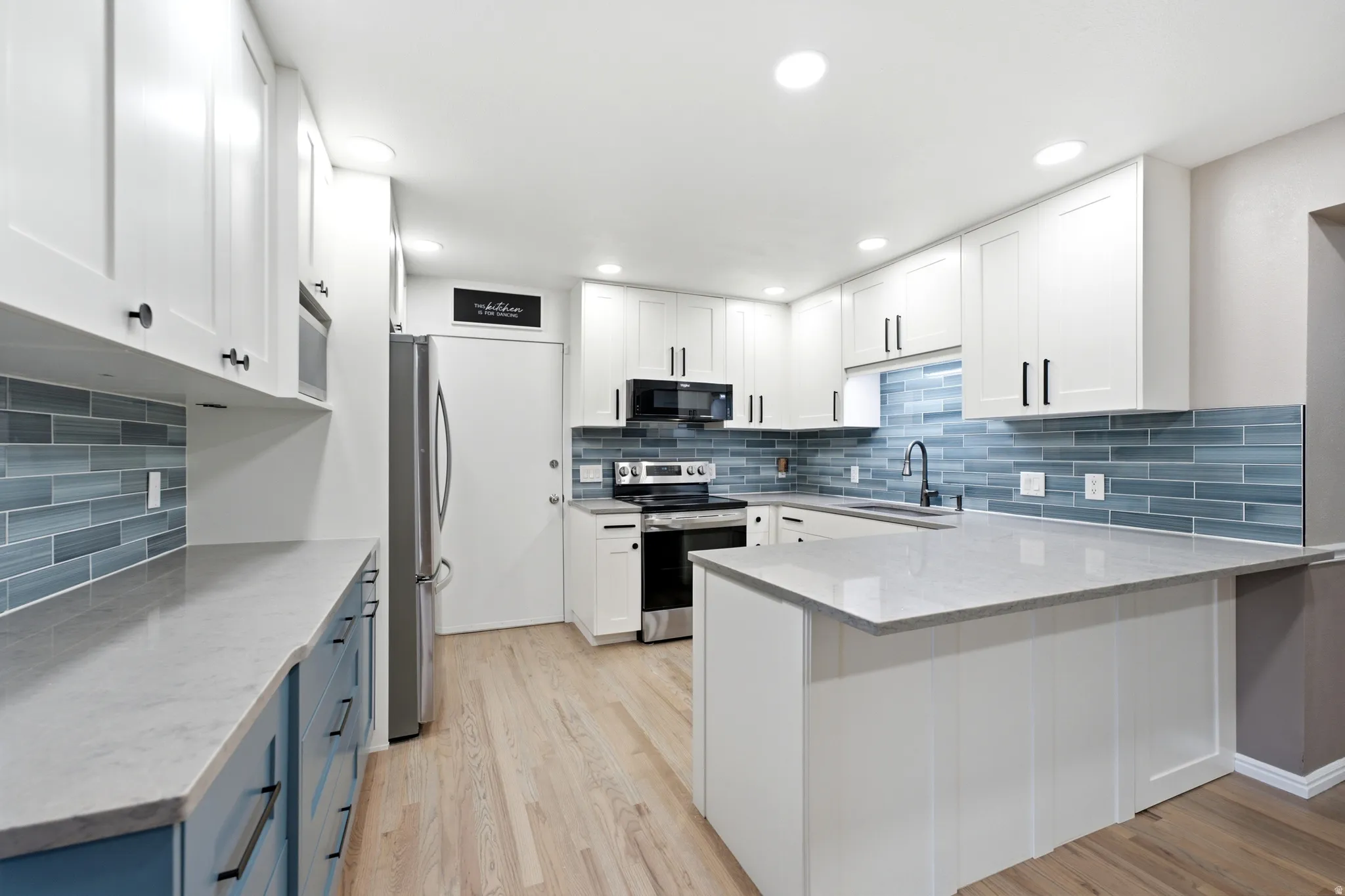 Light-toned wood-finish flooring, white cabinetry, integrated stainless steel appliances, light-colored countertops, and a blue tile backsplash