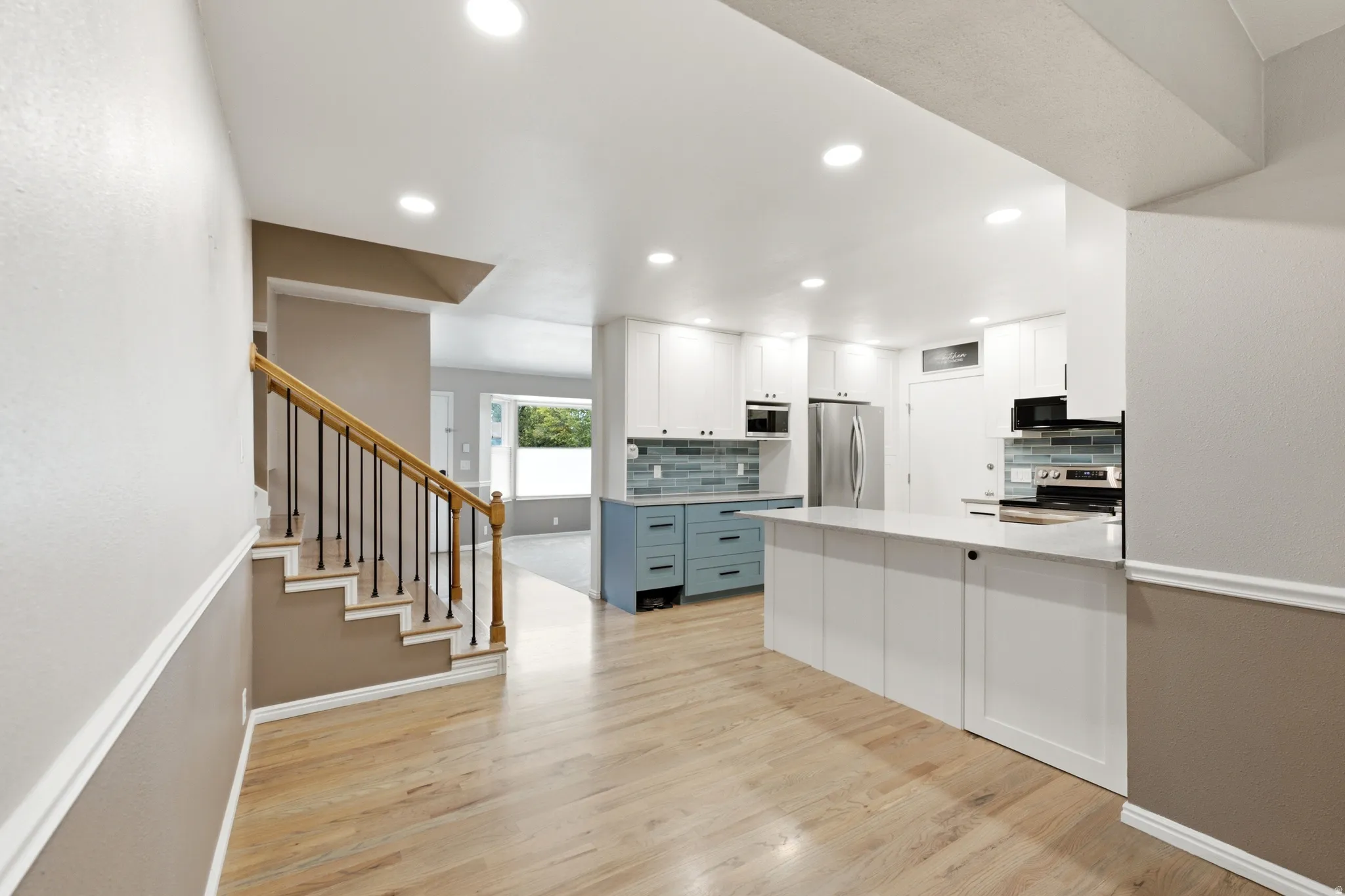 Recessed lighting illuminates the wood-finish flooring, two-tone kitchen cabinetry, stainless steel appliances, and a tiled backsplash
