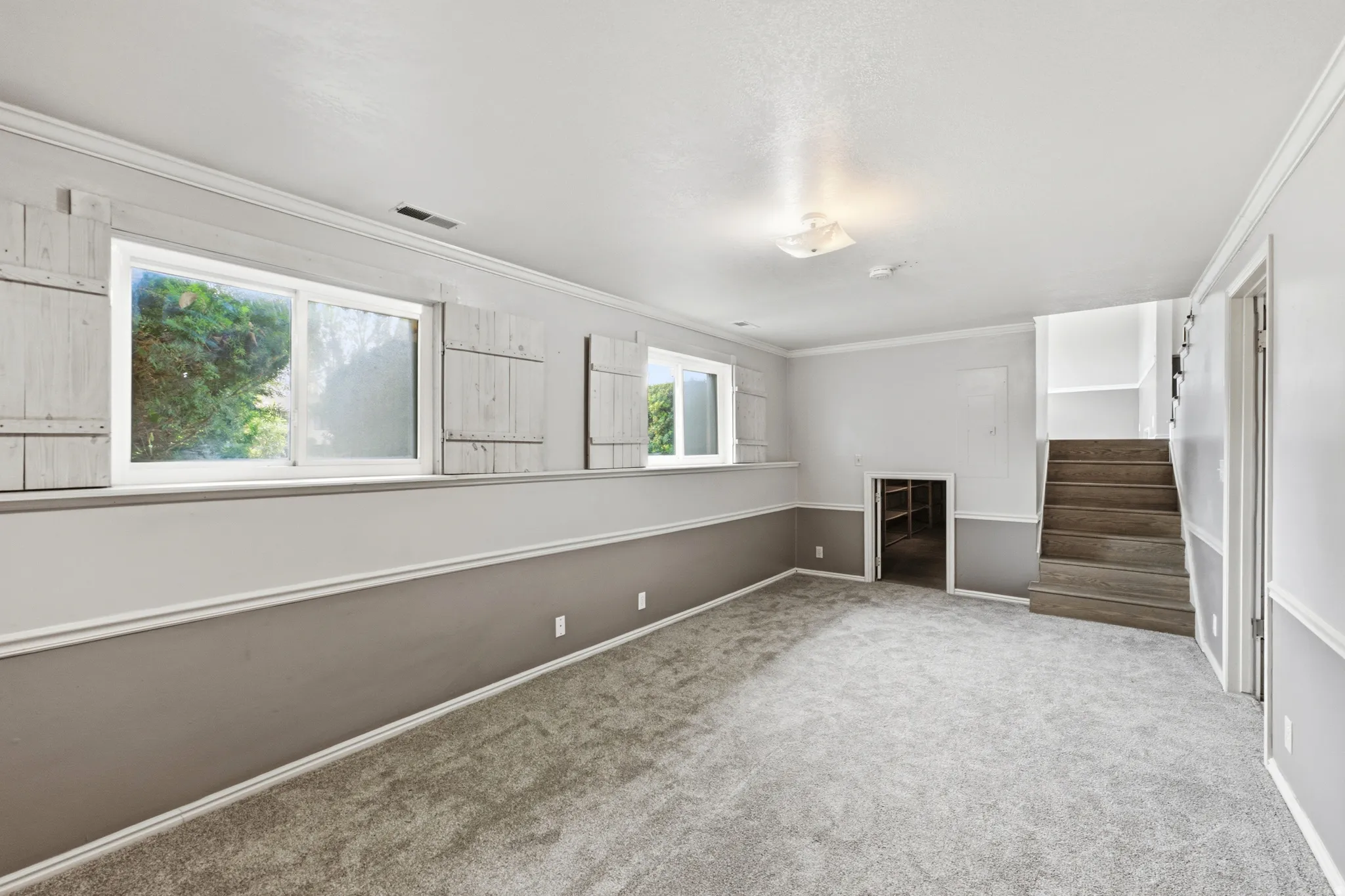 Spacious room featuring light gray carpet, dual windows with decorative shutters, a two-tone wall finish with chair rail molding, and an integrated staircase with wood-finish treads