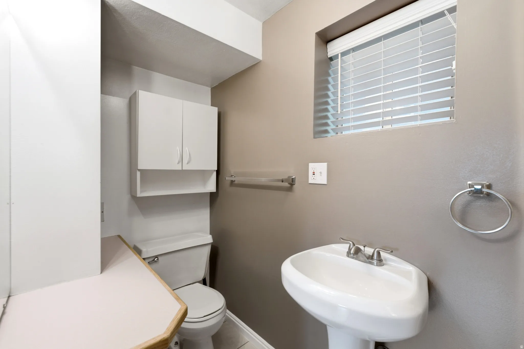 Pedestal sink with chrome fixtures, window with blinds, wall-mounted storage cabinet, and neutral wall paint