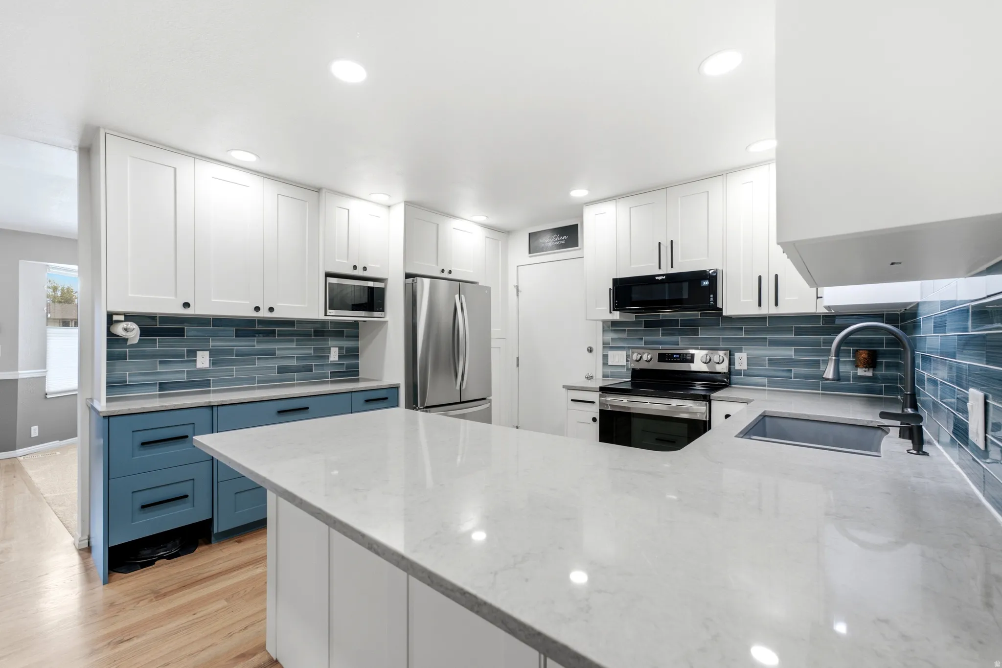 Contemporary kitchen featuring a large island with a light-toned countertop, white upper cabinetry, blue lower cabinetry, a blue glass tile backsplash, and stainless steel appliances