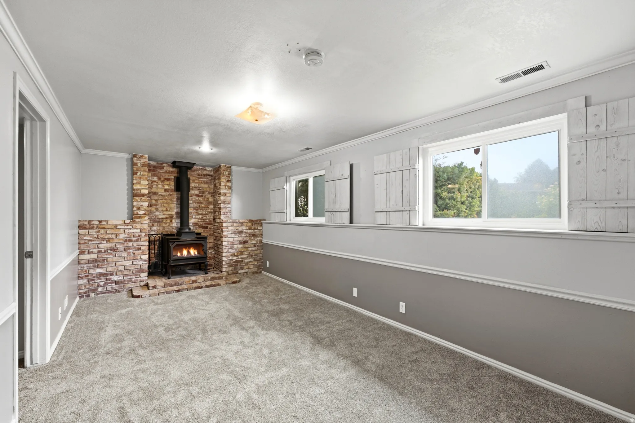 Wood-burning stove set within a brick surround, light gray carpeting, chair rail molding, and multiple windows with decorative shutters