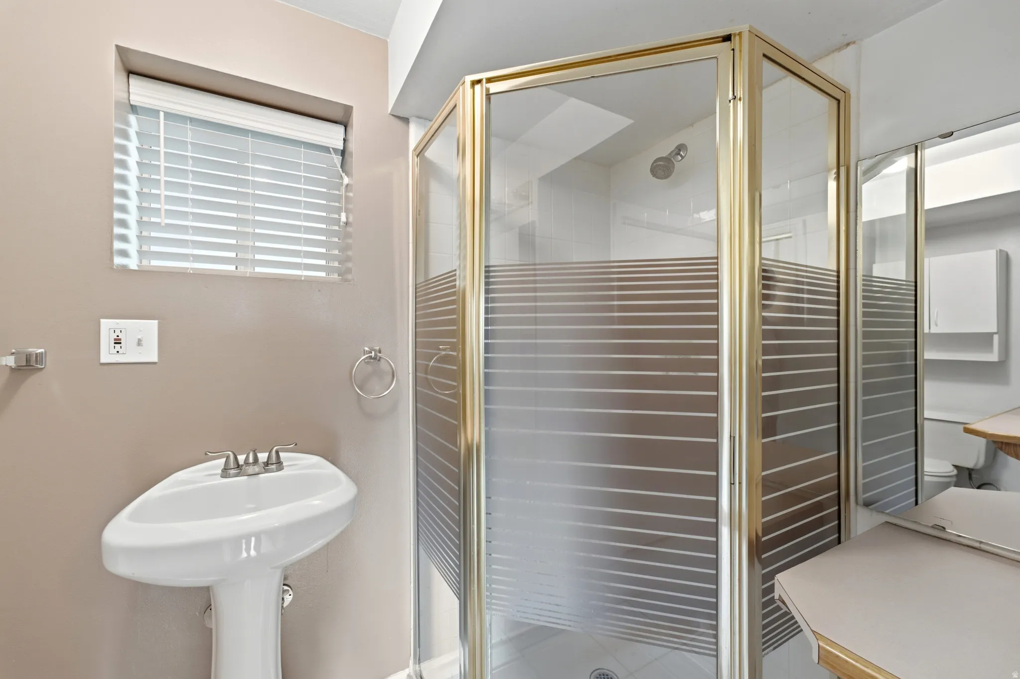 Pedestal sink with dual-handle faucet, window with horizontal blinds, and a gold-finish shower enclosure with patterned privacy glass