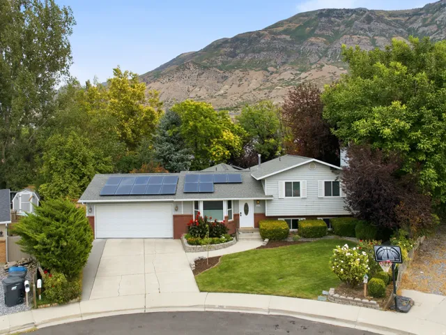 Light gray siding with brick accents, attached garage, solar panels, concrete driveway, and established landscaping