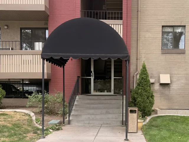Building exterior featuring brick and stucco finishes, balconies with vertical slat railings, a ground-level entrance with a black fabric awning, and concrete stairs