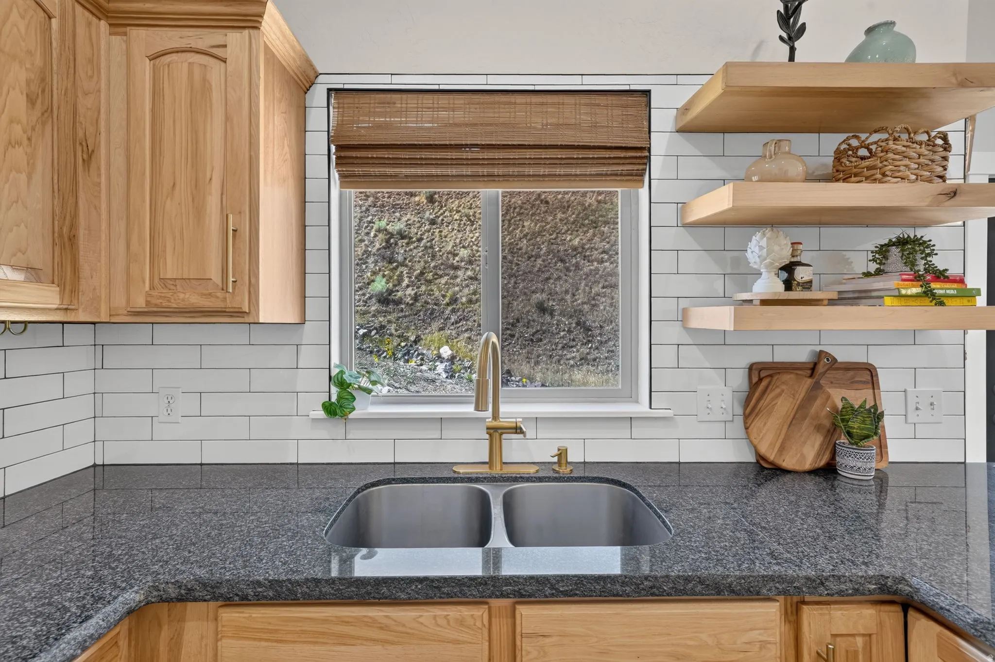 Dual basin stainless steel sink set within a dark speckled countertop, complemented by a white subway tile backsplash, light wood cabinetry, and open shelving