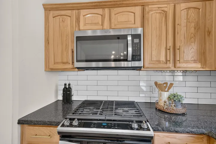 Light wood-finish cabinetry, stainless steel over-the-range microwave, subway tile backsplash, dark stone-finish countertop, and stainless steel gas range