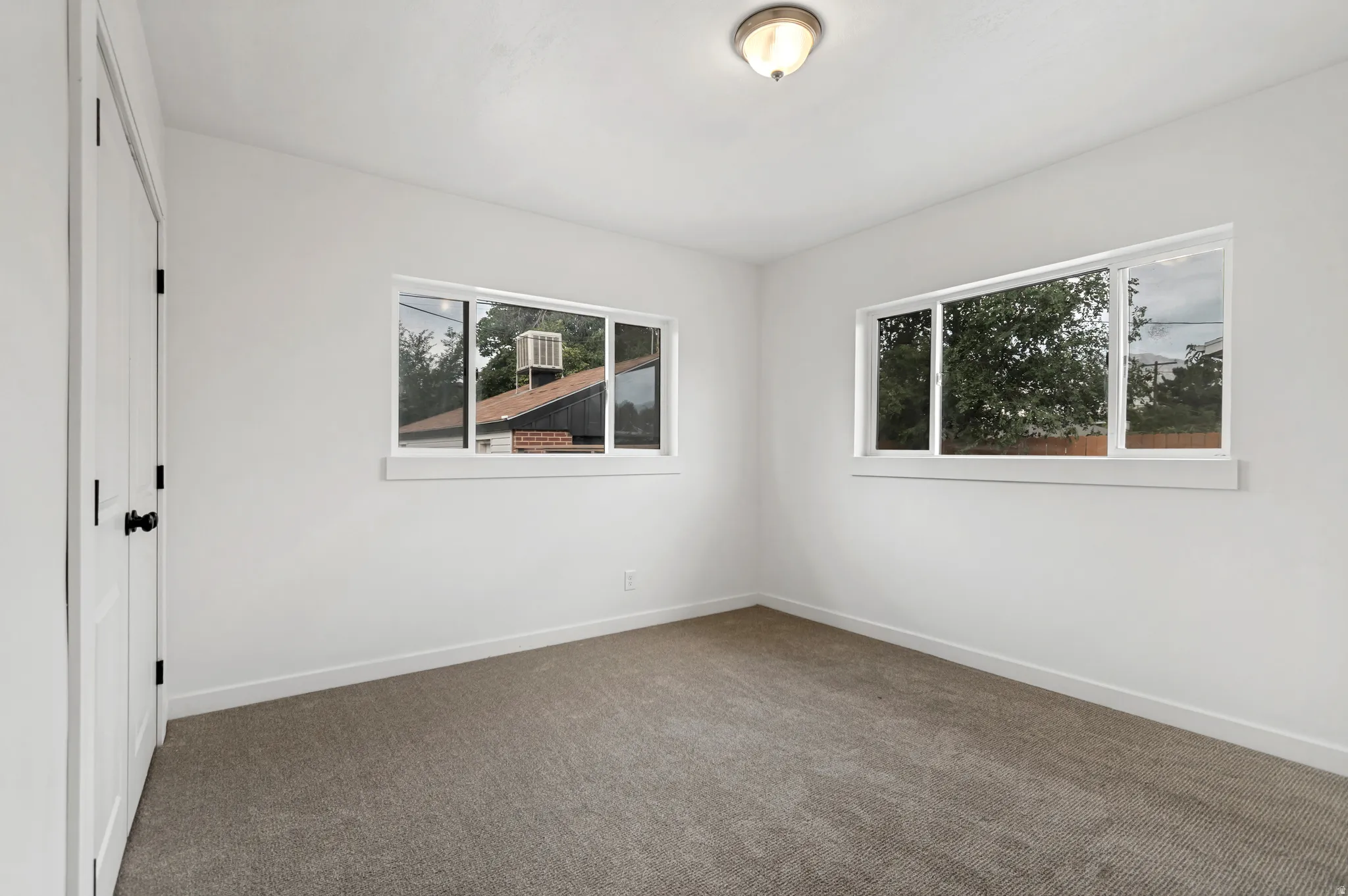 Neutral wall paint, ceiling-mounted light fixture, two windows with white trim, gray carpet flooring, and white trim baseboards