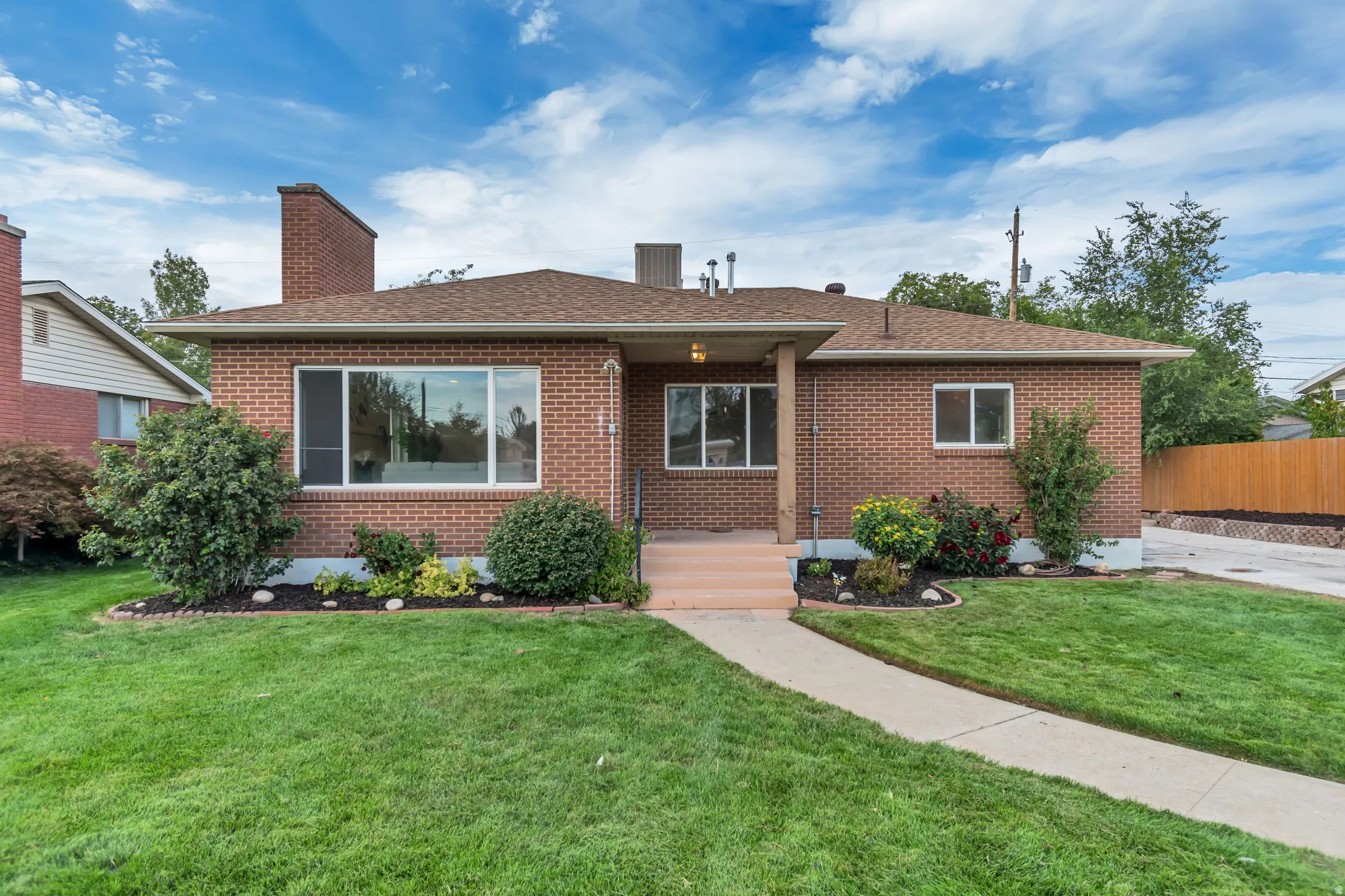 Brick exterior with a brown shingle roof, concrete pathway, and landscaped garden beds