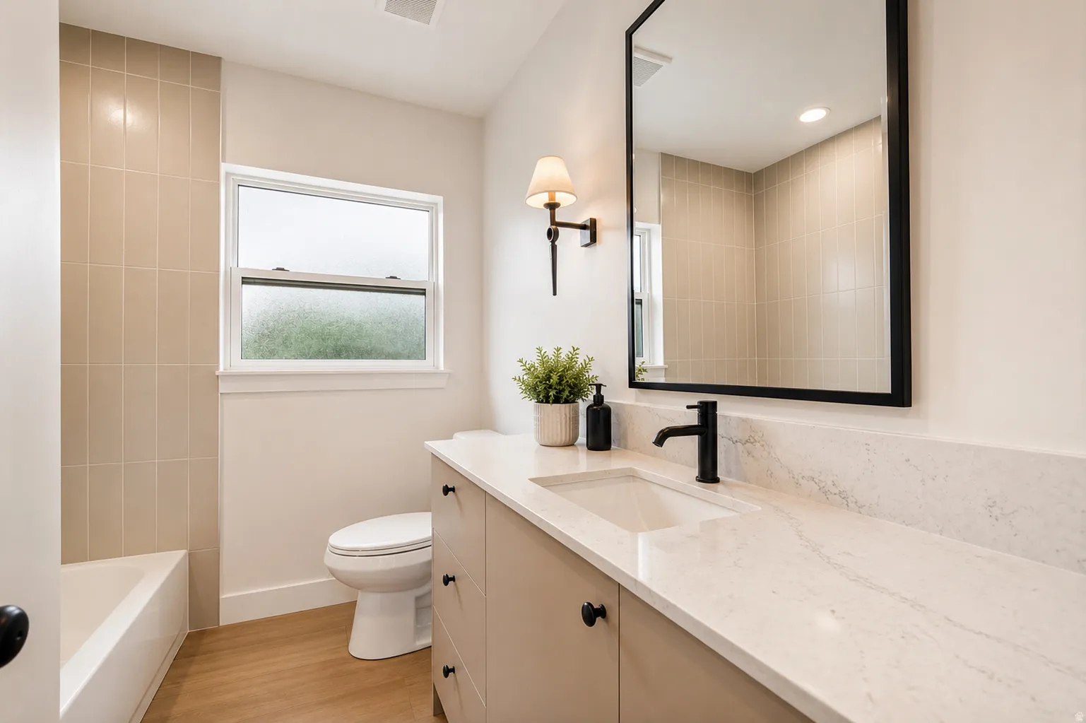 Wood-finish flooring, built-in bathtub with tiled surround, single-pane window, vanity with integrated sink and stone countertop, and a wall-mounted light fixture