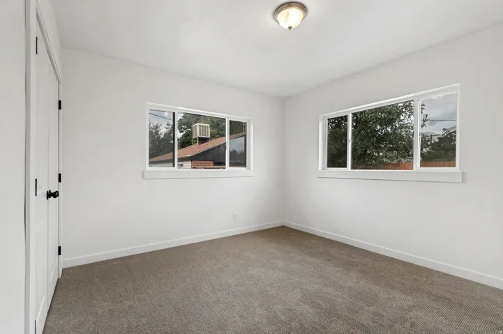 Neutral wall paint, ceiling-mounted light fixture, two windows with white trim, gray carpet flooring, and white trim baseboards