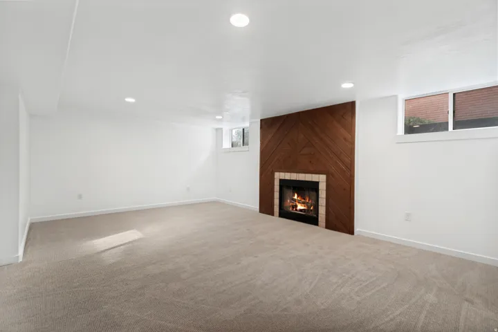 Recessed lighting illuminates a spacious room featuring a fireplace with a wood-paneled accent wall, a tiled hearth, and carpet flooring