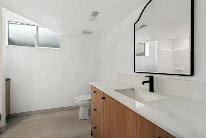 Light-toned wood-finish vanity with a single basin sink, white countertop, and matte black faucet