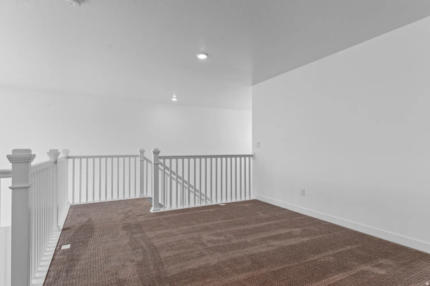 Spacious upper-level loft featuring wood-finish flooring, white railing with metal balusters, recessed ceiling lights, and white walls providing a bright, open atmosphere