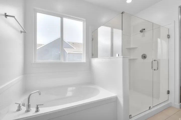 Deep soaking tub with polished chrome fixtures, glass-enclosed shower with built-in shelving, white tiled walls, and recessed lighting