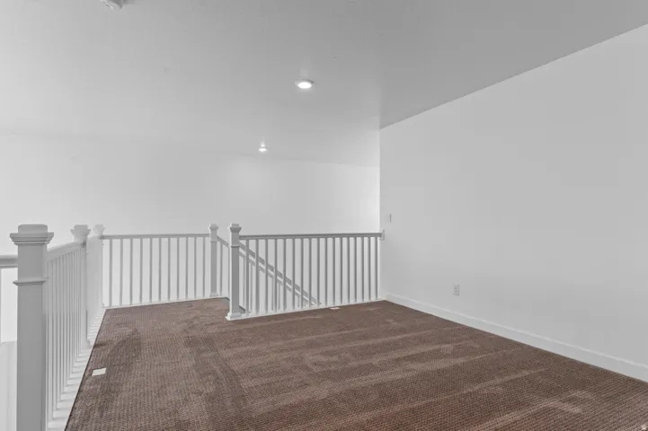 Spacious upper-level loft featuring wood-finish flooring, white railing with metal balusters, recessed ceiling lights, and white walls providing a bright, open atmosphere