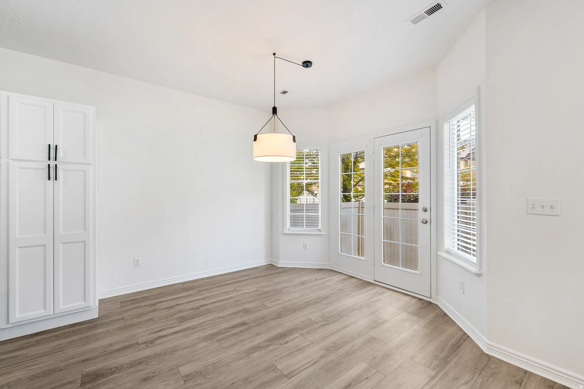 Wood-finish flooring, white cabinetry with black hardware, modern pendant lighting, multiple windows with blinds, and a glass-paneled door
