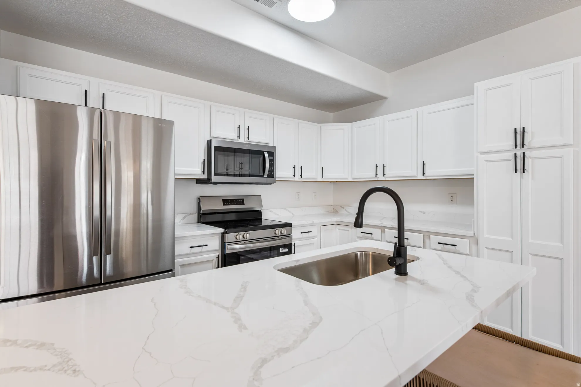 Kitchen featuring white cabinetry, stainless steel appliances, a kitchen island with an undermount sink, a matte black faucet, and white countertops