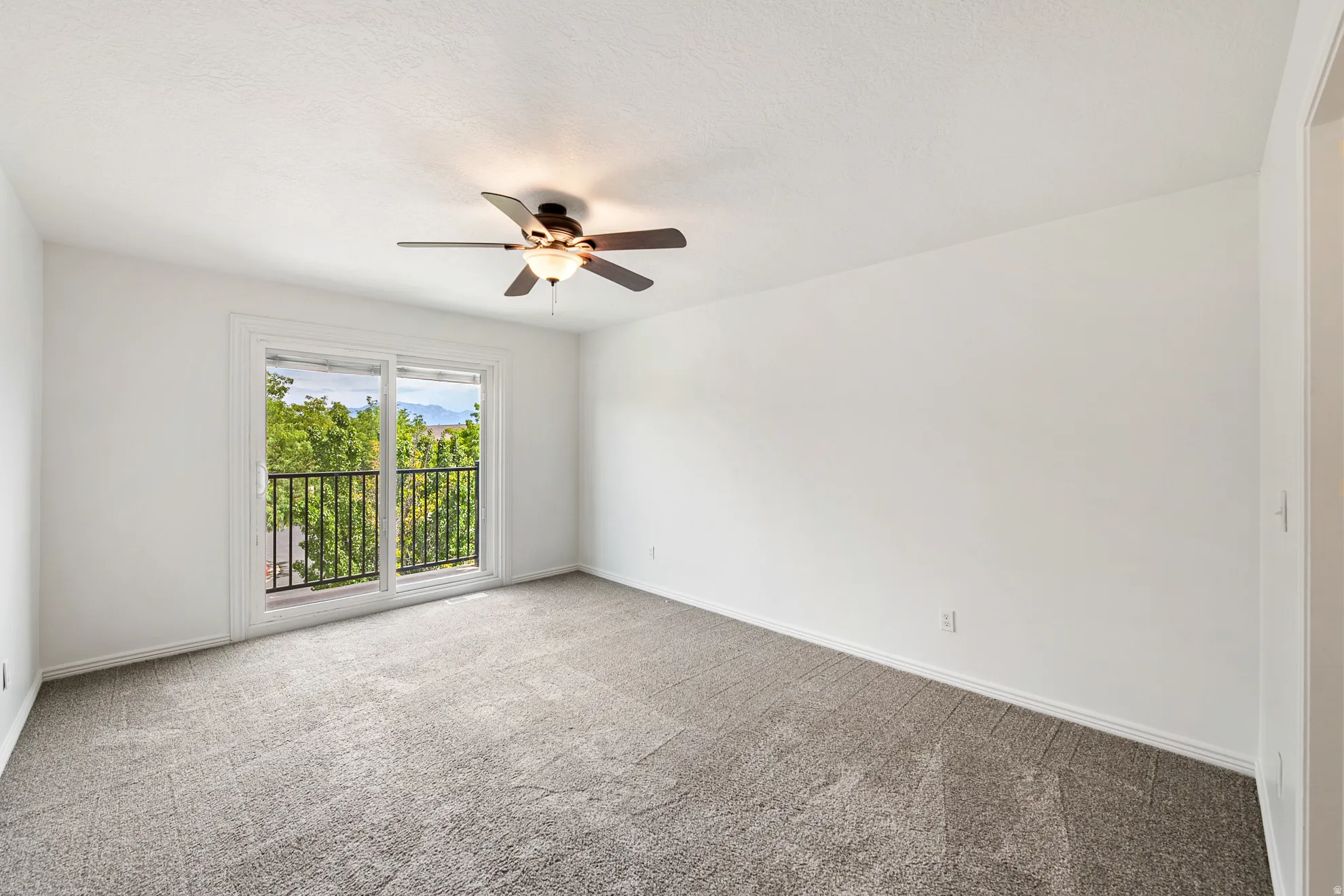 Sliding glass door leading to a balcony with black wrought iron railing, ceiling fan with light fixture, and neutral carpeting