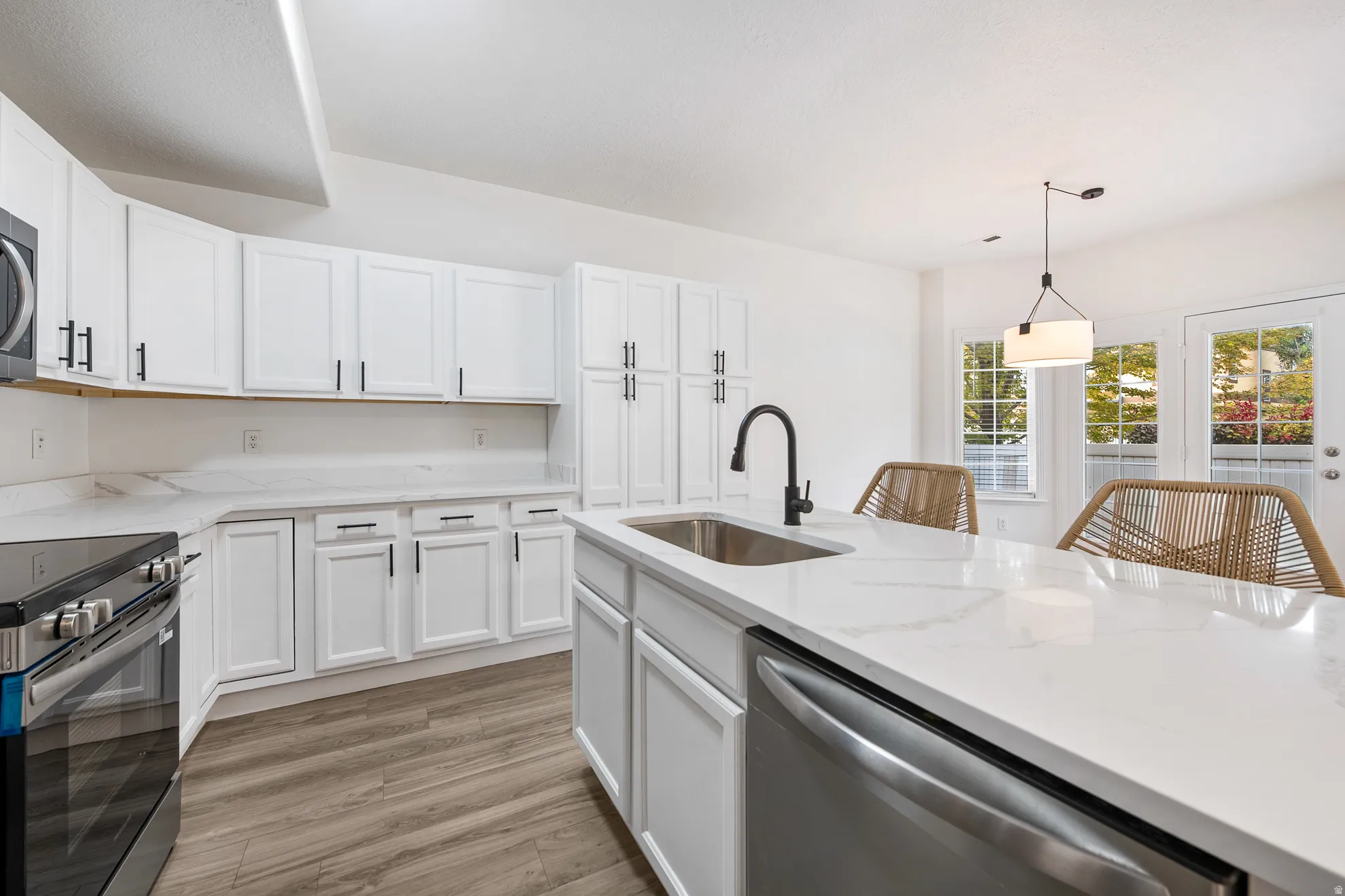 White shaker cabinetry with black hardware, wood-finish flooring, stainless steel appliances, a kitchen island with sink, and white countertop surfaces