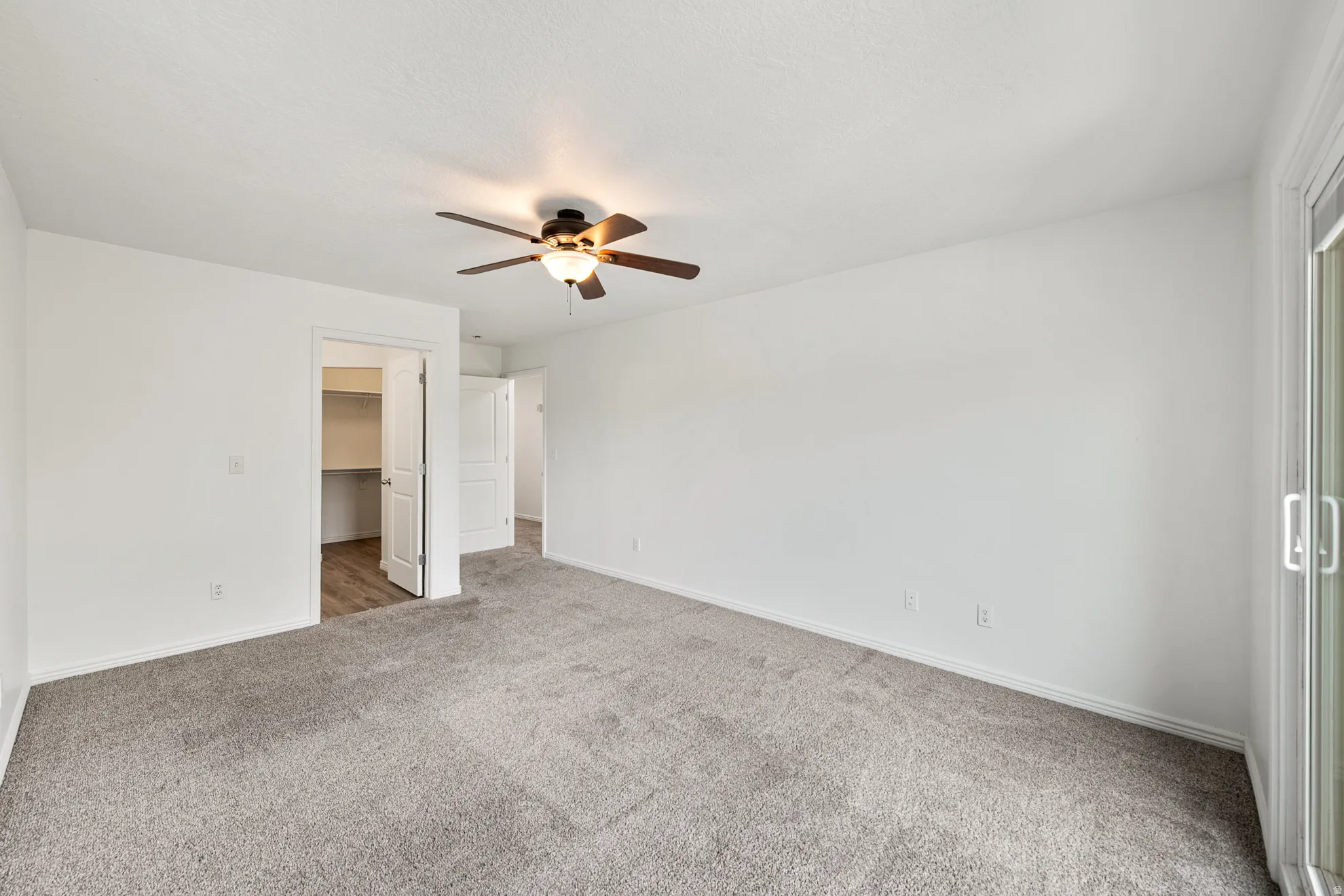 Spacious room featuring light gray carpeting, crisp white walls, a ceiling fan with integrated lighting, and a sliding glass door