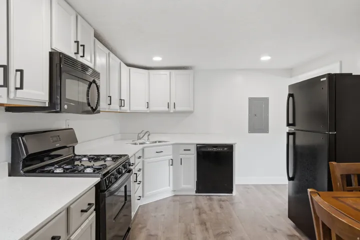 Modern kitchen featuring white cabinetry with hardware accents, stainless steel gas range and range hood, black dishwasher, black refrigerator with bottom freezer, white subway tile backsplash, and wood-finish flooring throughout the open layout