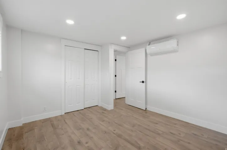 Recessed ceiling lighting, wood-finish flooring, white sliding closet doors, white paneled door with black hardware, and a wall-mounted climate control unit