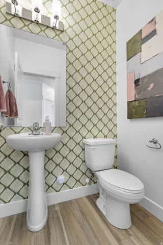 Pedestal sink with chrome faucet, toilet, wood-finish flooring, and wall-mounted vanity lighting