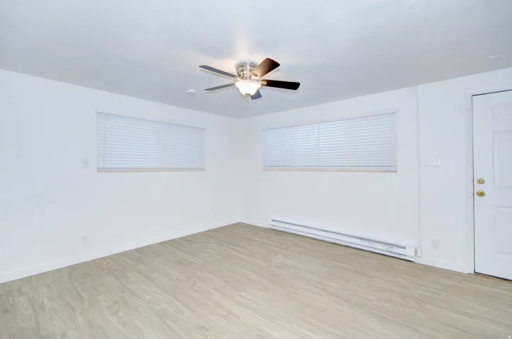 Spacious interior featuring wood-finish flooring, two windows with blinds, a ceiling fan with integrated lighting, and a white paneled door