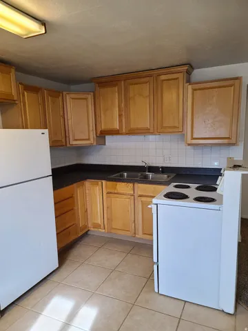 Kitchen featuring wood-finish cabinetry, a double basin stainless steel sink, tiled backsplash, white refrigerator, and a white electric range