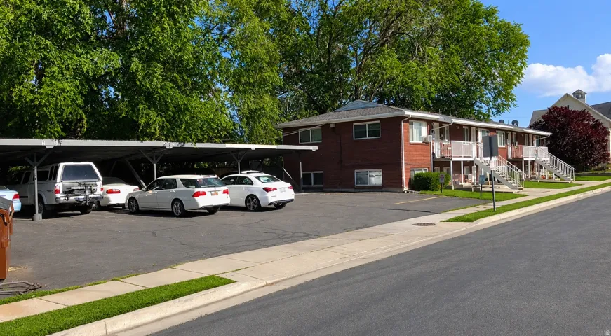 Brick exterior building with multiple units, covered parking structure, paved parking area, and mature trees
