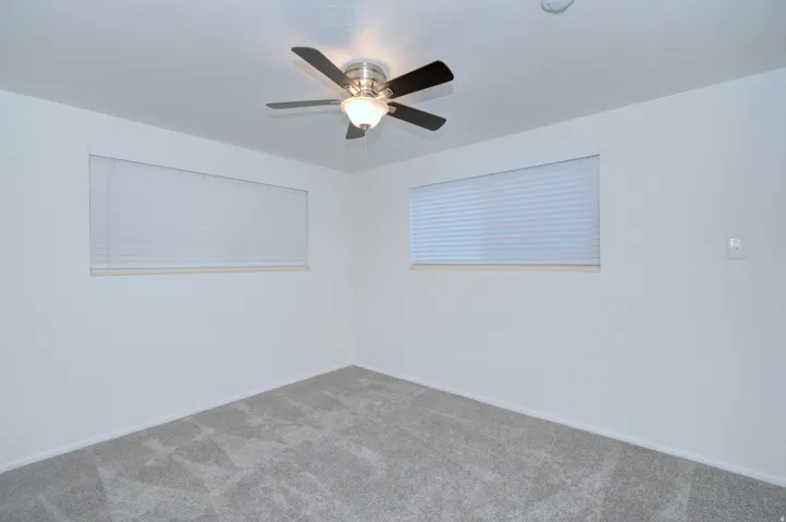 Light gray carpet flooring, white walls, two windows with horizontal blinds, and a ceiling fan with light fixture