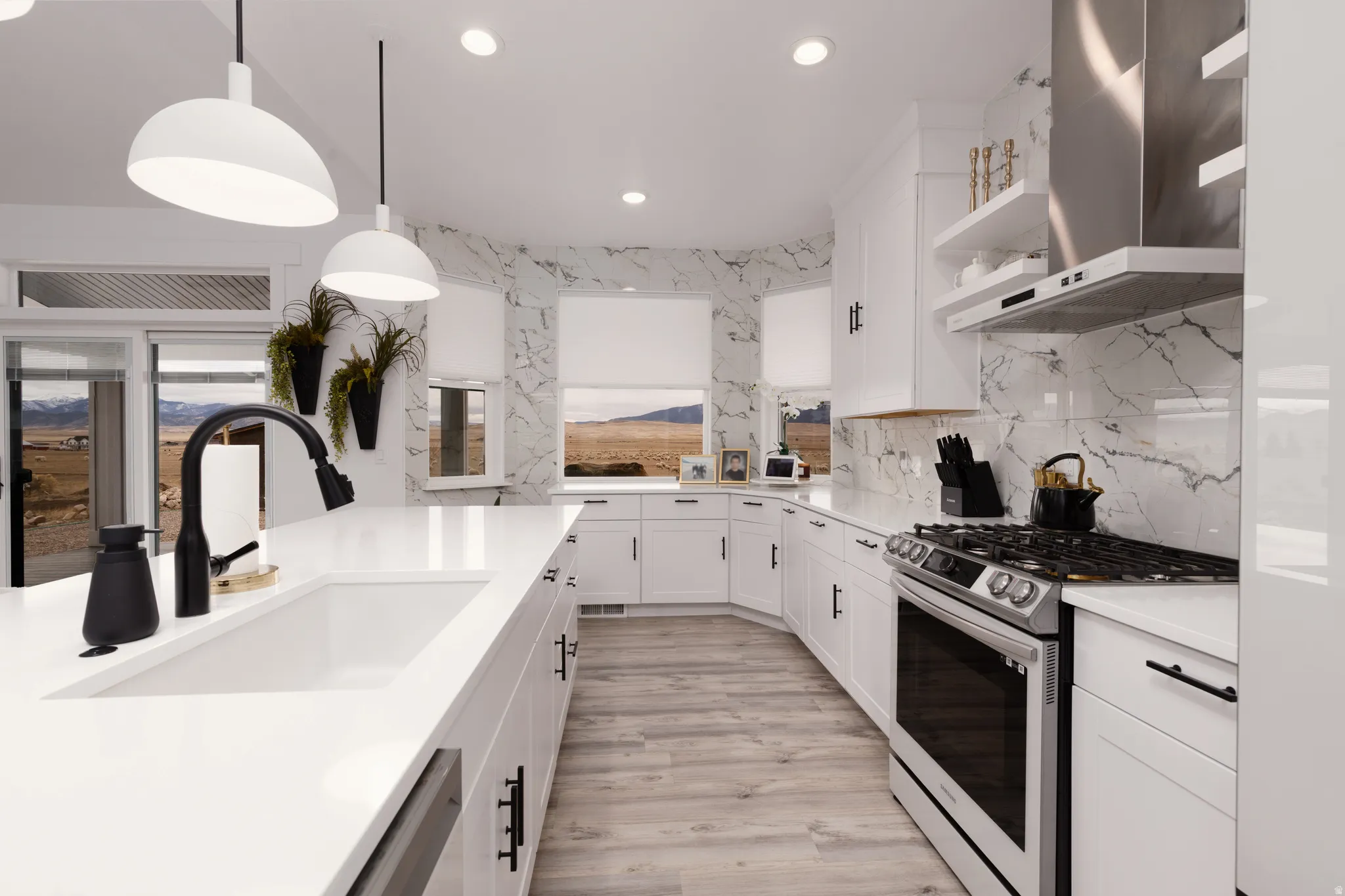 White cabinetry with black hardware, integrated stainless steel appliances, large kitchen island with single basin sink and black faucet, light wood-finish flooring, and white marble-patterned backsplash