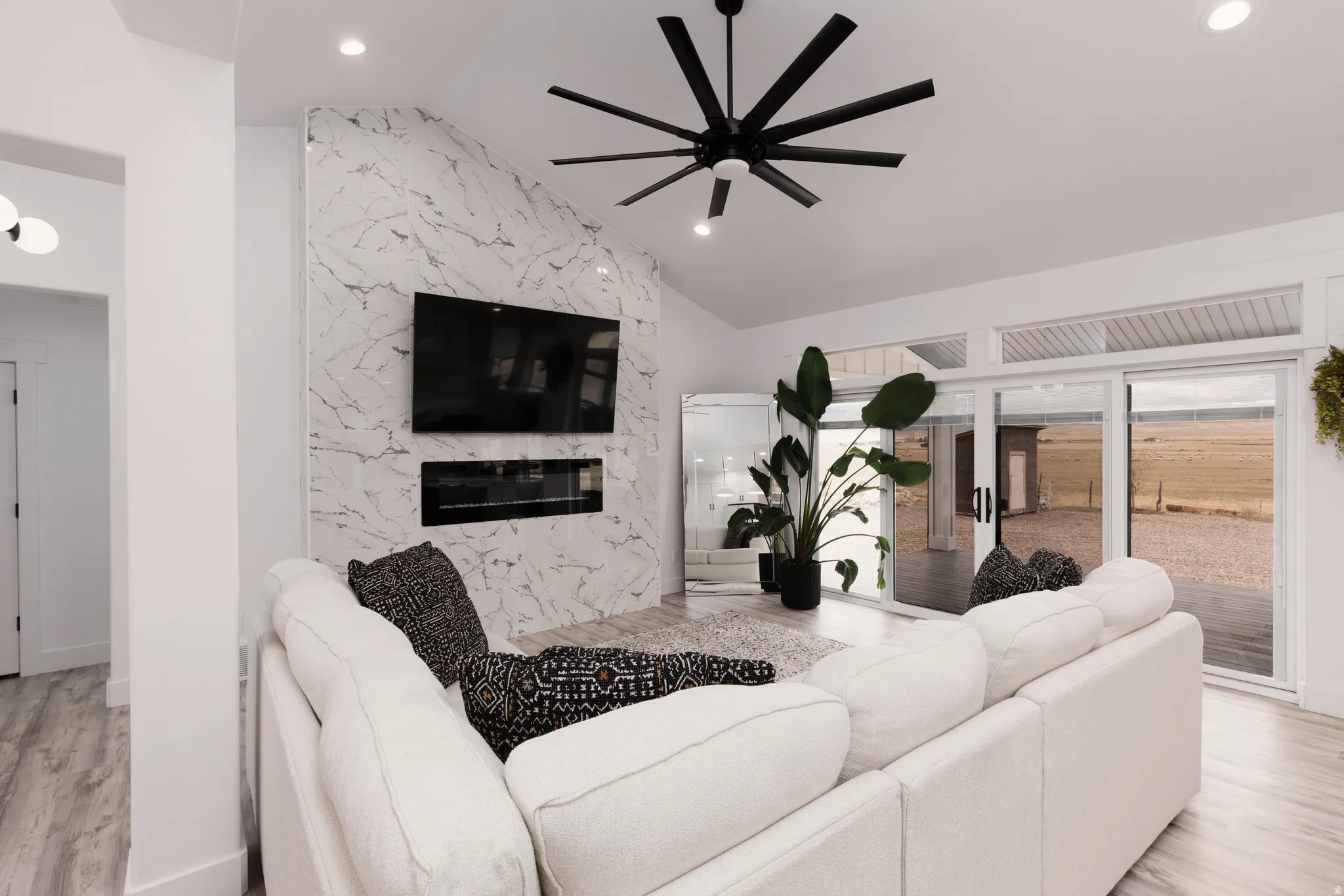 Modern living room showcasing a curved cream sectional, white marble fireplace surround with dual built-in fireplaces, wall-mounted flat-screen television, black modern ceiling fan with integrated recessed lighting, light wood-finish flooring, and sliding