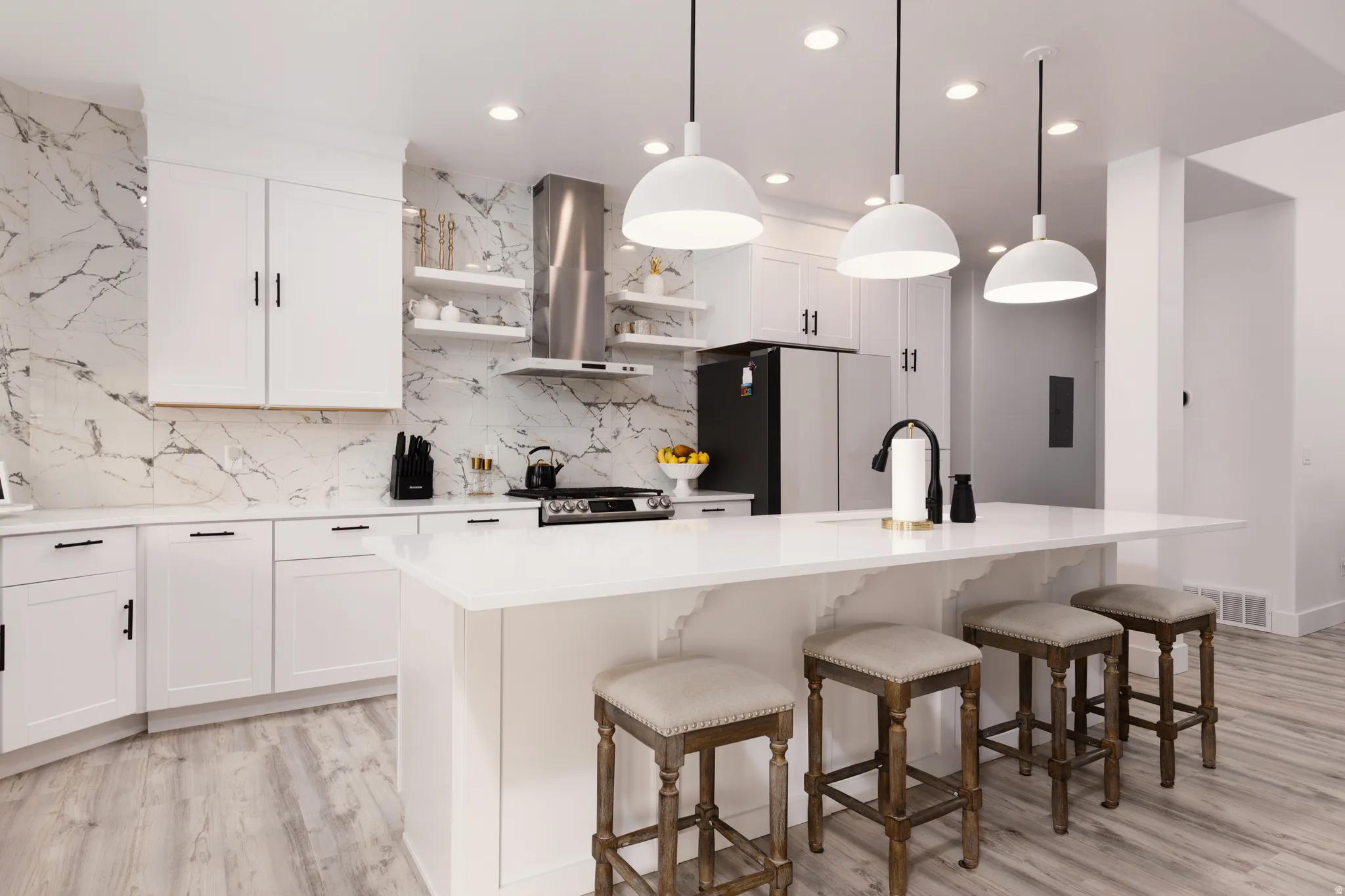 White cabinetry, patterned backsplash, stainless steel range hood, white island with seating, and light wood-finish flooring