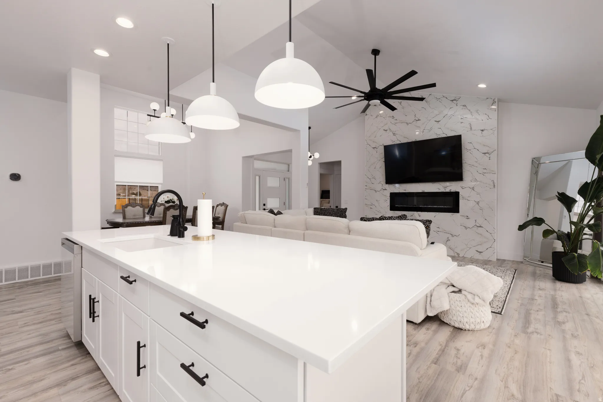 Open-concept living space featuring a kitchen island with white cabinetry and dark hardware, pendant lighting, wood-finish flooring, and a fireplace with a stone-finish surround